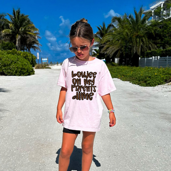 Boujee On My Parent's Dime DTF Heat Transfer, product image showing a young girl wearing a pink t-shirt with the slogan 'boujee on my parents dime' printed on it, outdoors on a sunny beach day.
