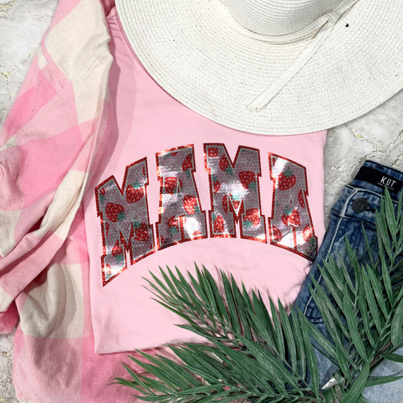 Mama with Strawberries Sequin Transfer, pink t-shirt with strawberry-themed sequin design, casual summer outfit with a wide-brim hat, denim shorts, and a plaid shirt, laid on a marble background with green palm leaves.