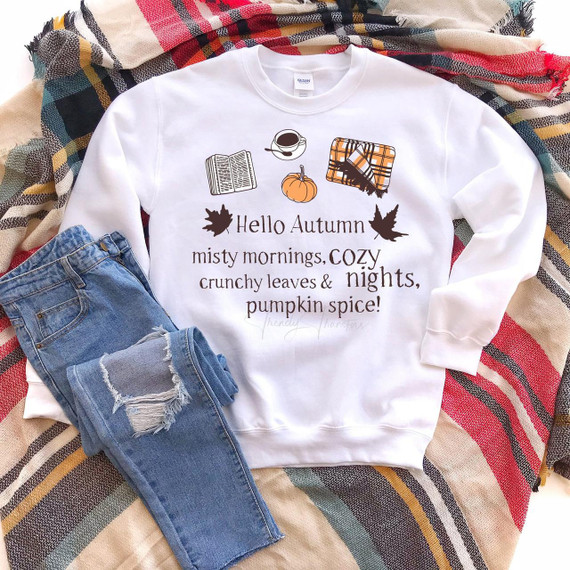Hello Autumn Sublimation Transfer, a white sweatshirt with autumn-themed designs and text, featuring a pumpkin, a book, a coffee cup, a plaid blanket, and text that says 'Hello Autumn misty mornings, cozy crunchy leaves & nights, pumpkin spice!' placed on a multicolored striped blanket with a pair of ripped blue jeans.