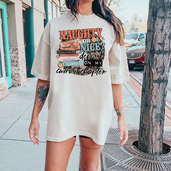 Naughty Or Nice? Depends On My Chapter DTF Heat Transfer, a woman wearing a beige oversized t-shirt with a colorful book-themed graphic and text, standing outdoors on a sidewalk.