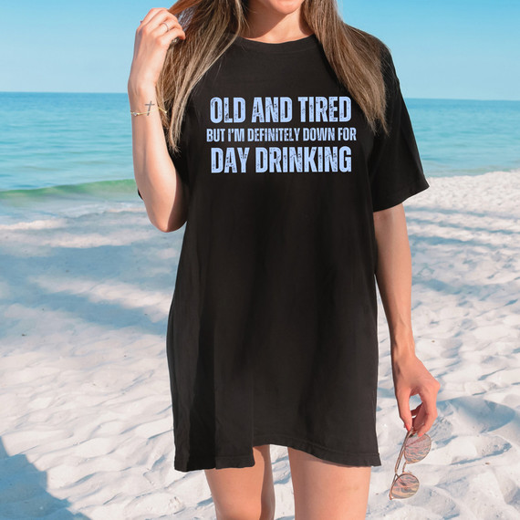 Old And Tired But I'm Definitely Down For Day Drinking DTF Heat Transfer, a woman on a beach wearing a black T-shirt with the humorous slogan in blue text.