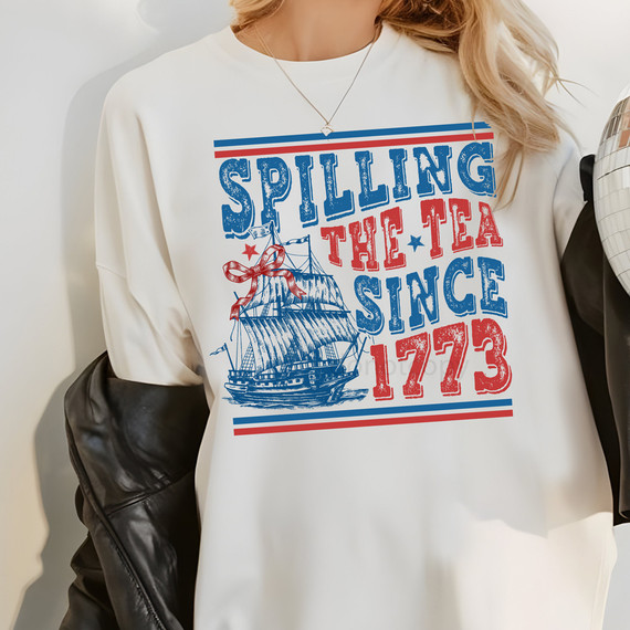 Distressed Spilling The Tea Since 1773 DTF Heat Transfer, a white sweatshirt featuring a vintage sailing ship graphic and patriotic red and blue text design.