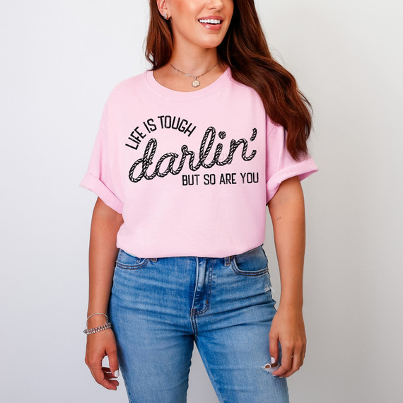Life Is Tough Darlin' But So Are You DTF Heat Transfer, a woman wearing a pink t-shirt with black text, casual style with rolled-up sleeves and ripped jeans.