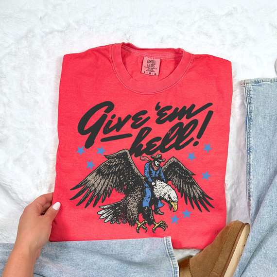 Give 'Em Hell Eagle DTF Heat Transfer, vibrant red graphic T-shirt featuring a fierce eagle with a man riding on its back, accompanied by blue stars and bold black text.