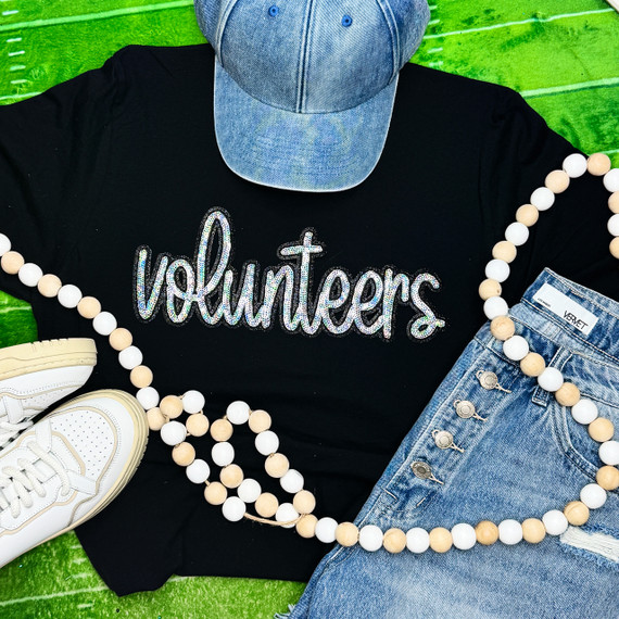 Volunteers Sequin Patch, glittering embroidered patch on black t-shirt with the word 'volunteers' in cursive, surrounded by casual denim clothing and a beaded necklace, placed on a vibrant green surface.