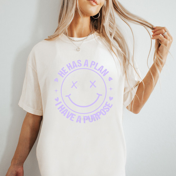 He Has A Plan I Have A Purpose Happy Face Lavender DTF Heat Transfer, image of a woman wearing a white t-shirt with a lavender heat transfer design of a smiling face with crossed eyes and the text "He Has A Plan I Have A Purpose" around it.