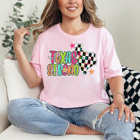 Texas Strong Checkered State DTF Heat Transfer, a colorful and vibrant heat transfer design featuring the state of Texas in a checkered pattern with rainbow colors and stars, on a pink t-shirt worn by a woman.