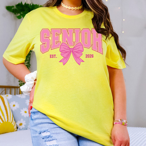 Senior 2026 Pink Bow DTF Heat Transfer, pink text with bow design on a yellow T-shirt worn by a person.