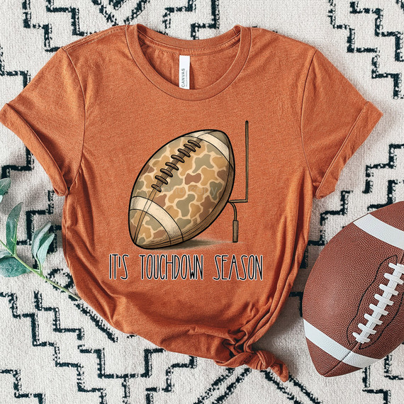 It's Touchdown Season Camo Football DTF Heat Transfer, a graphic of a football with camouflage pattern and the text 'It's Touchdown Season' on a burnt orange t-shirt.