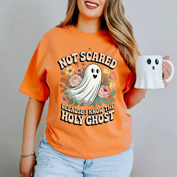 Not Scared Because I Know The Holy Ghost Retro DTF Heat Transfer, a woman wearing an orange T-shirt with a ghost graphic and floral design, holding a white mug with ghost face.