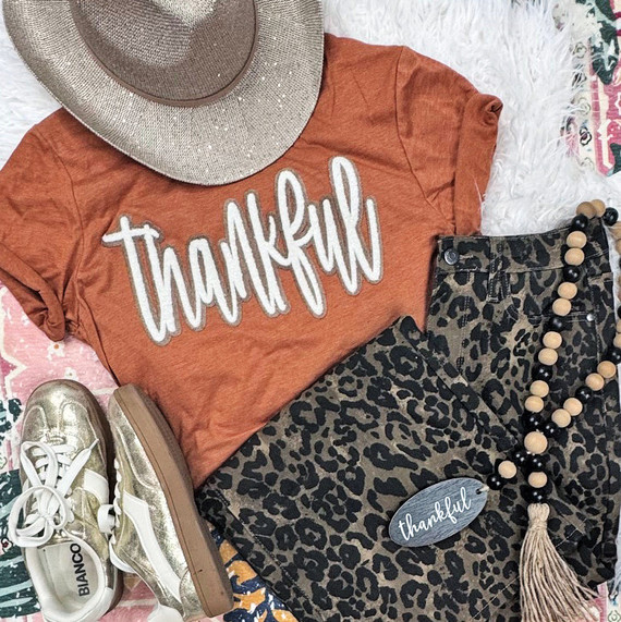 Double Layer Stacked Thankful Fluffy Patch, fashion outfit with a rust-colored T-shirt with 'thankful' in white embroidered letters, leopard print pants, gold sneakers, a beige sunhat, and a beaded necklace.