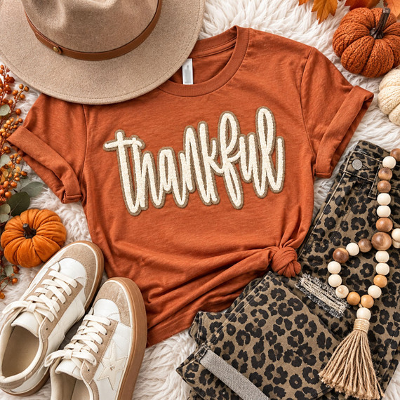 Double Layer Stacked Thankful Fluffy Patch, fashion outfit with a rust-colored T-shirt with 'thankful' in white embroidered letters, leopard print pants, gold sneakers, a beige sunhat, and a beaded necklace.