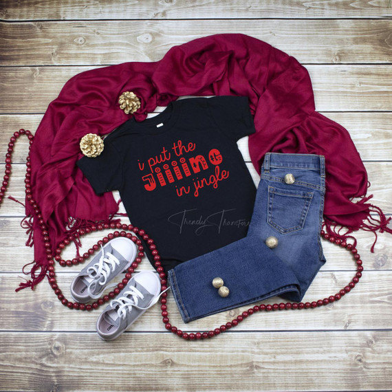 Jiiiing in Jingle FINAL STOCK Screen Print Transfer, a black t-shirt with festive red text and ornaments design, paired with jeans, sneakers, a red scarf, and holiday accessories on a wooden background.