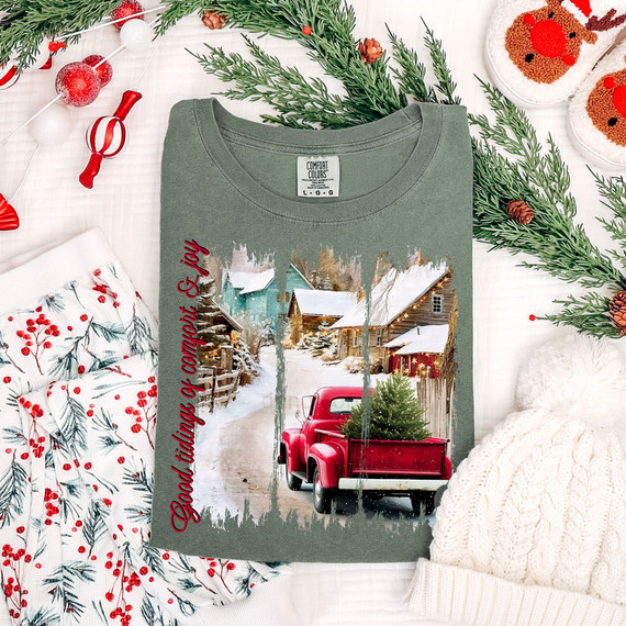 Good Tidings Of Comfort & Joy DTF Heat Transfer, a Christmas-themed t-shirt with a winter scene of a snowy village and a red truck carrying a Christmas tree, surrounded by holiday decorations.