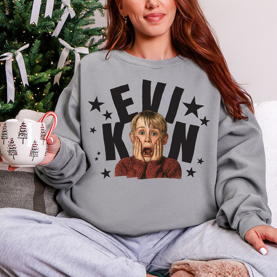 Kevin Stars BLACK DTF Heat Transfer, woman wearing a sweatshirt with a surprised boy graphic and text, holding a Christmas themed mug, sitting in front of a decorated Christmas tree.