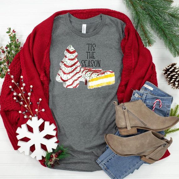 Tis' The Season Christmas Cake Screen Print Heat Transfer, festive graphic on a grey t-shirt with Christmas tree and cake designs, surrounded by holiday decor and clothing items.
