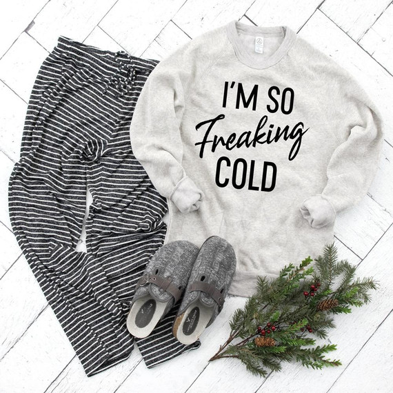 I'm So Freaking Cold Screen Print Transfer, cozy sweatshirt with black lettering, striped pajama pants, gray slippers, and holiday greenery on white wooden background.