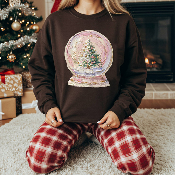 Faux Hand Painted Tree Snow Globe DTF Heat Transfer, Christmas snow globe with a decorated tree inside, on a person wearing a dark sweater and red plaid pajama pants, near a decorated Christmas tree and fireplace.