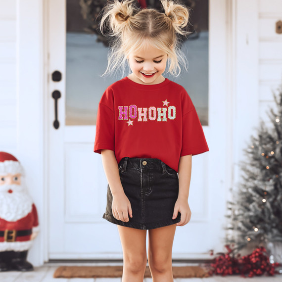 Faux Glitter Ho Ho Ho DTF Heat Transfer, image of a young girl smiling and wearing a red t-shirt with glittery Ho Ho Ho design on front, in festive Christmas setting.