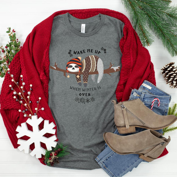 Wake Me When Winter Is Over Sloth Screen Print Heat Transfer, a graphic T-shirt featuring a cute sloth wearing winter attire and hanging from a tree branch, surrounded by holiday plants and accessories on a white background.