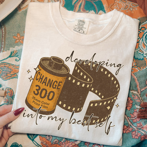 Developing Into My Best Self DTF Heat Transfer, white t-shirt with a design of a film reel and a film canister labeled 'CHANGE 300' for 35mm print film, emphasizing self-growth and creativity.