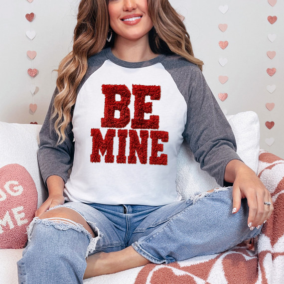 Faux Fluffy Be Mine DTF Heat Transfer, image of a woman wearing a white and gray baseball-style shirt with red fluffy 'BE MINE' text, sitting on a cozy sofa with Valentine's Day heart decorations in the background.