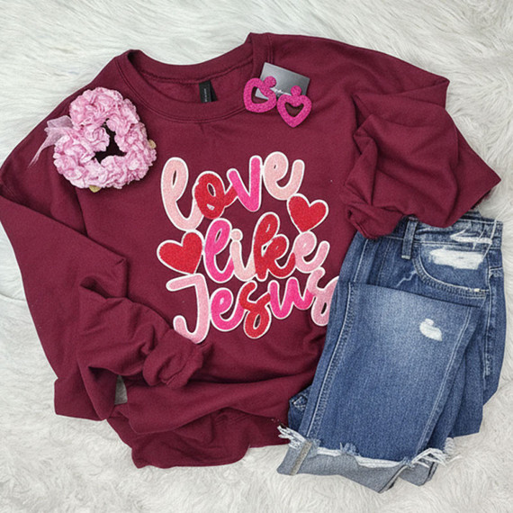 Love Like Jesus Multicolor Chenille Patch, image of a burgundy sweatshirt with the text 'love like Jesus' in pink and white, decorated with red hearts, accompanied by pink heart-shaped earrings and a pink floral scrunchie, paired with distressed jeans.