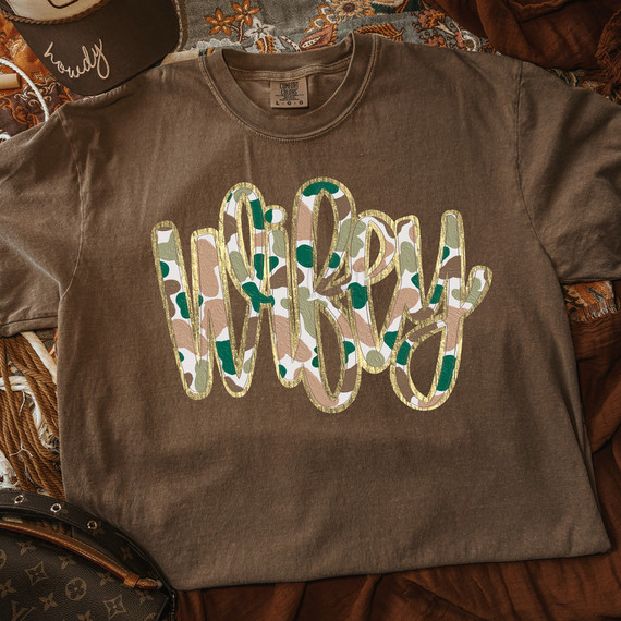 Wifey Script Camo Faux Gold Metallic DTF Heat Transfer, photo of a brown sweatshirt featuring a camo print hello script design with faux gold metallic finish, laid out on a patterned fabric surface.