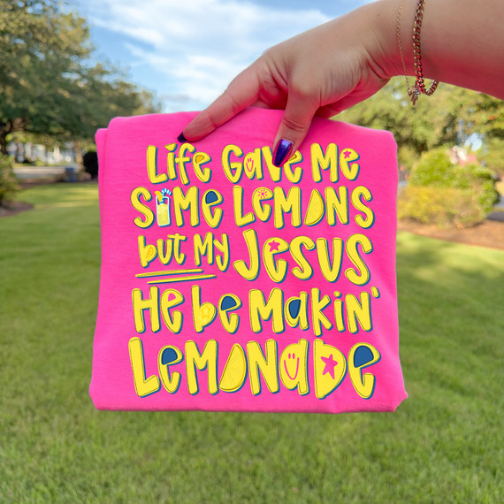 Life Gave Me Some Lemons, But My Jesus He Be Makin’ Lemonade DTF Heat Transfer, vibrant pink fabric with colorful, playful text and lemon graphics in yellow, blue, and pink, held outdoors in a park setting.