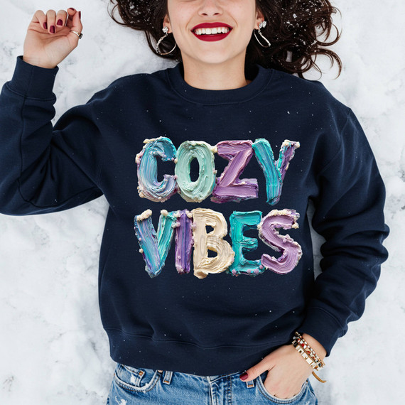 Faux Painted Cozy Vibes DTF Heat Transfer, colorful painted text design on a navy sweatshirt worn by a smiling woman with curly hair.