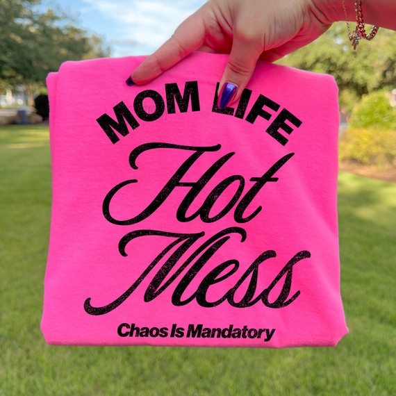 Distressed Mom Life Hot Mess – Chaos Is Mandatory BLACK DTF Heat Transfer, pink fabric with black text design and a person's hand holding the fabric outdoors.