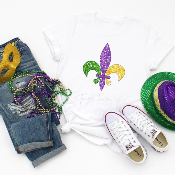 Glitter Effect Fleur De Lis Mardi Gras Sublimation Transfer, product image of a white t-shirt with a colorful glitter fleur de lis design in purple, green, and yellow, surrounded by Mardi Gras accessories like beads, a green hat with sequins, white sneakers, and a gold mask.