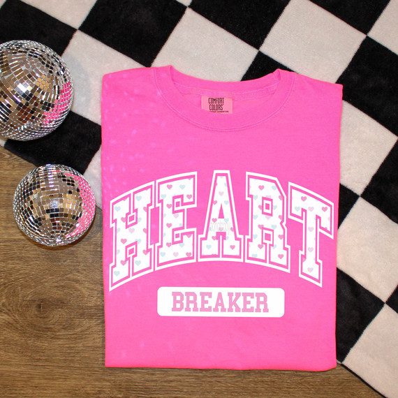Pastel Heart Breaker DTF Heat Transfer, pink T-shirt with Heart Breaker graphic design, printed with pastel-colored hearts, featuring a bold white font and outlined letters, placed on a wooden surface with black and white checkered background.