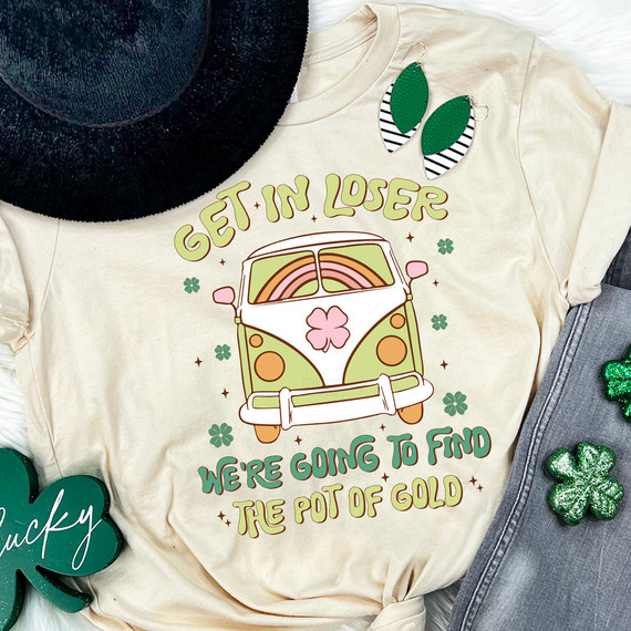 Get In Loser, We're Going to Find the Pot of Gold Retro Van DTF Heat Transfer on a white t-shirt, with green shamrock earrings, a black hat, and Saint Patrick's Day decorations around.