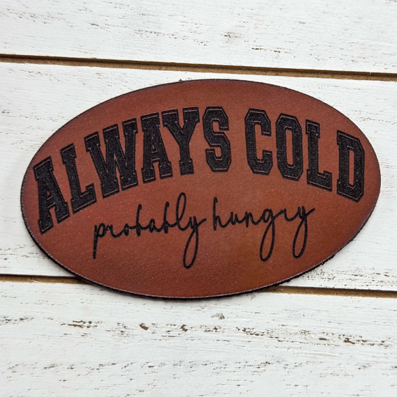 Always Cold Probably Hungry Leather Hat Patch, oval-shaped leather patch with black embroidered text 'ALWAYS COLD probably hungry' on a white wooden background.