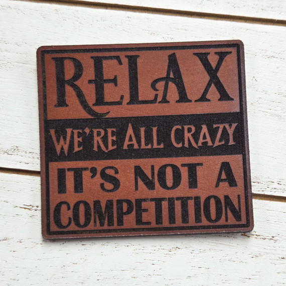 Relax We're All Crazy Leather Hat Patch, a brown leather patch with black text and design, saying 'Relax We're All Crazy It's Not a Competition'.