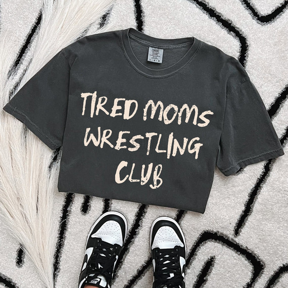 Tired Moms Wrestling Club, black graphic T-shirt with white distressed font text, casual wear for mothers involved in wrestling or fitness, laid flat on a textured rug with sneakers nearby.
