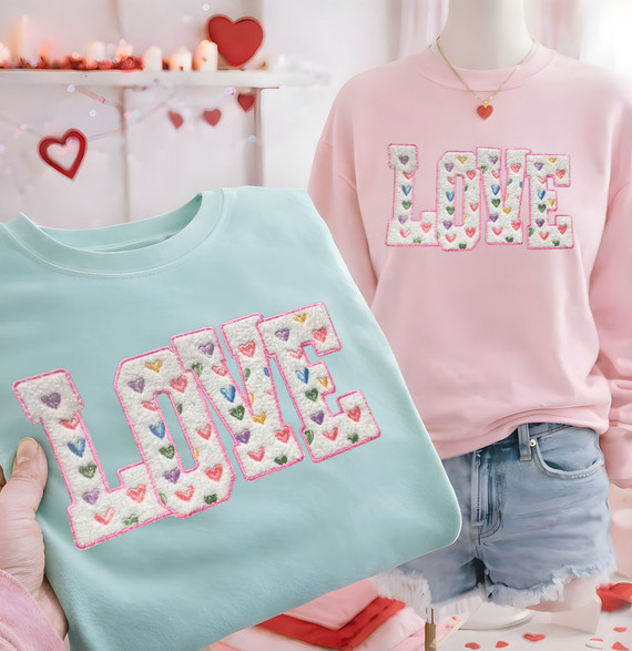 Fluffy white LOVE patch with multicolor embroidered hearts and pink stitched edging, shown applied to pastel sweatshirts for a Valentine’s themed boutique display.