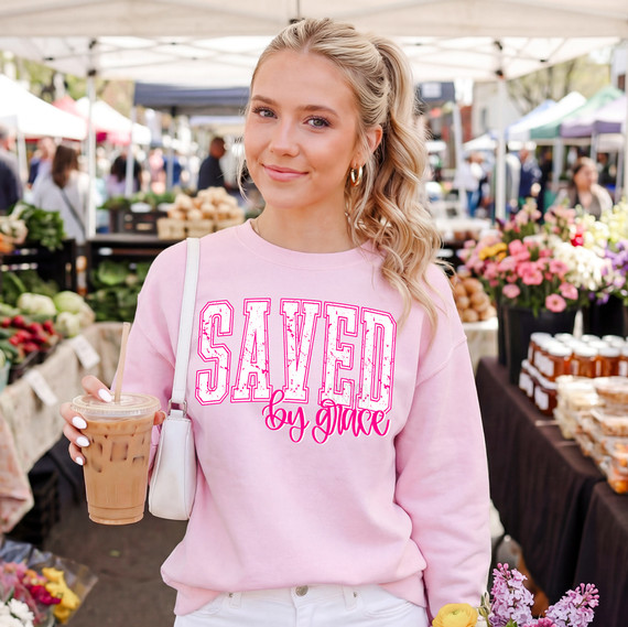 Distressed Saved By Grace DTF Heat Transfer, design on a pink sweatshirt worn by a woman at an outdoor market.