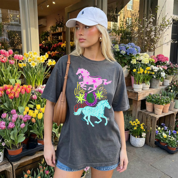 Y2K Horses DTF Heat Transfer, woman wearing a dark T-shirt with colorful unicorn and star designs, standing among vibrant spring flowers in a flower shop.