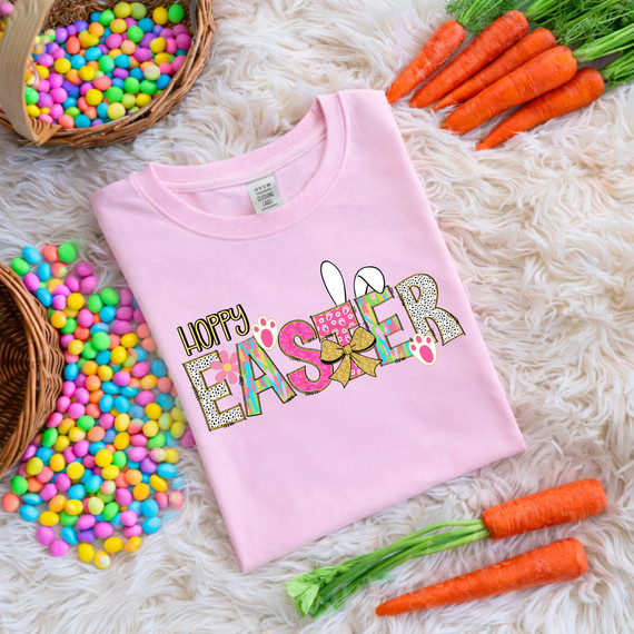 Hoppy Easter DTF Heat Transfer, pink t-shirt with Easter themed design featuring bunny ears, colorful letters, paw prints, and a bow, surrounded by carrots and colorful candies.