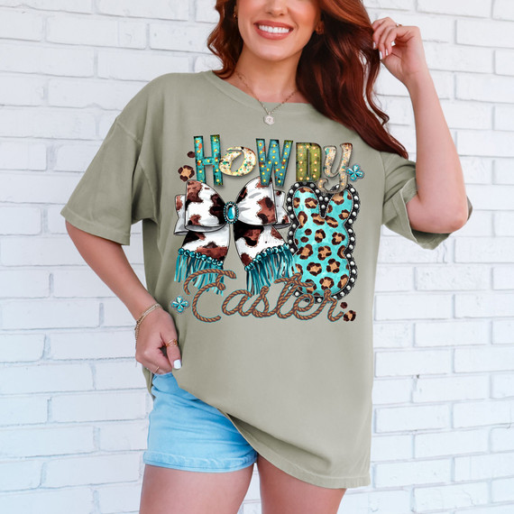Howdy Easter Western Bunny DTF Heat Transfer, woman wearing a green t-shirt with Easter and western-themed design, including a cow print bow and leopard print bunny, against a white brick wall.