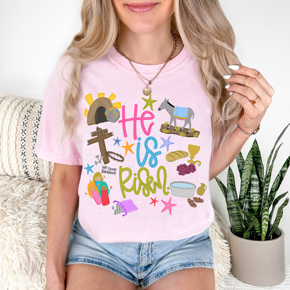 He Is Risen Icons DTF Heat Transfer, colorful religious and biblical-themed graphic design featuring a cross, rock with sun rays, donkey, bread, wine, water bowl, and flowers on a woman's pink T-shirt.