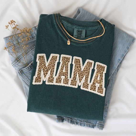 Faux Embroidered Mama Leopard DTF Heat Transfer, close-up of a dark green T-shirt with leopard print 'MAMA' text embroidery, folded on light denim jeans with dried flowers in the background.