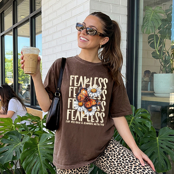 Fearless Butterfly DTF Heat Transfer, woman wearing a brown t-shirt with butterfly and flower design, holding iced coffee, outdoor cafe scene.