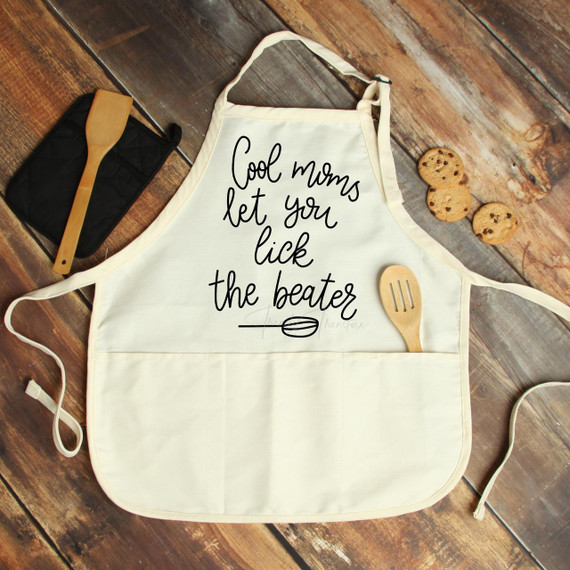 Product Cool moms let you lick the beater Sublimation Transfer, an apron with the humorous phrase printed on it, placed on a rustic wooden surface with a spatula, cookies, and baking tools around it.