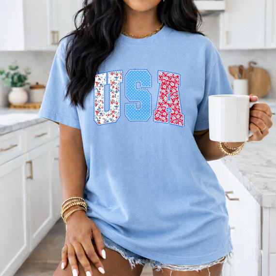 USA Patchwork DTF Heat Transfer, woman wearing a blue T-shirt with patchwork USA letters in floral and polka dot patterns, holding a white mug in a modern kitchen.