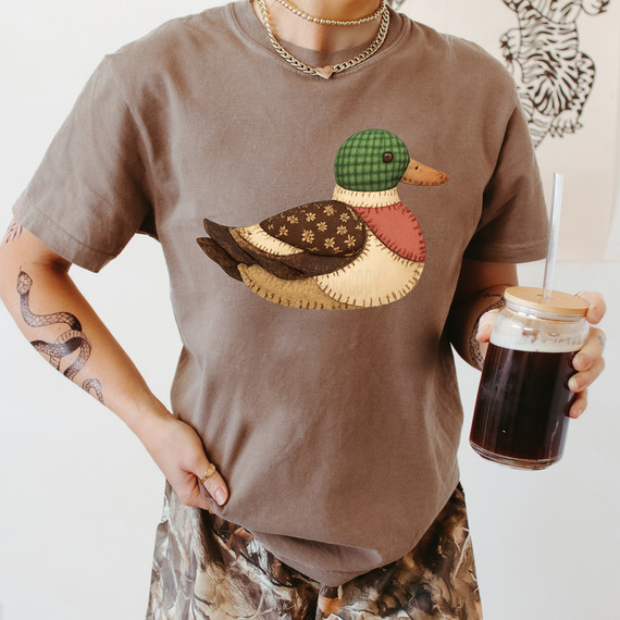 Faux Fabric Patchwork Mallard Duck DTF Heat Transfer, close-up of a person wearing a brown T-shirt with a patchwork duck design, holding a glass of iced coffee