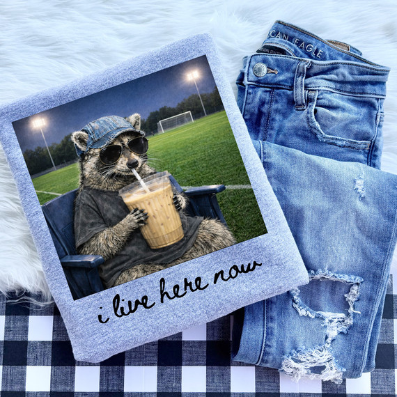 I Live Here Now Soccer DTF Heat Transfer, a humorous print of a raccoon wearing sunglasses and a denim cap, drinking bubble tea on a soccer field background, designed for custom apparel.