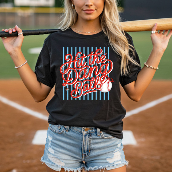 Hit The Dang Ball Script DTF Heat Transfer, a baseball-themed graphic design on a black t-shirt worn by a woman holding a baseball bat on a baseball field.